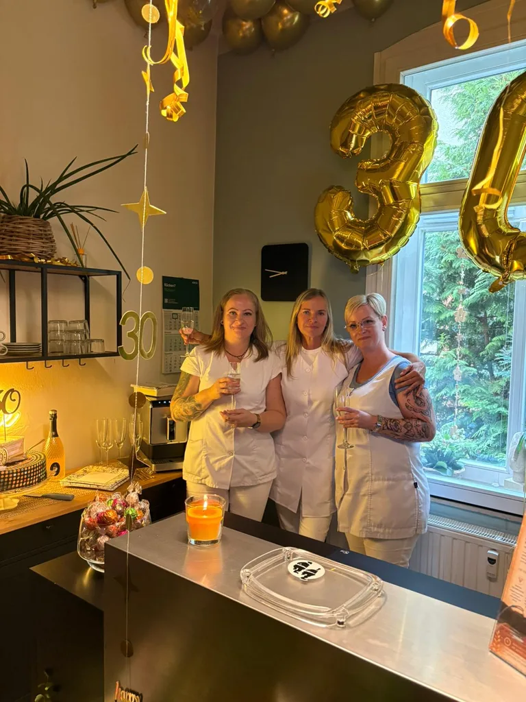 Drei Mitarbeiterinnen des Physiotrom-Teams in weißer Arbeitskleidung stoßen unter goldenen 30-Luftballons mit Sekt an.